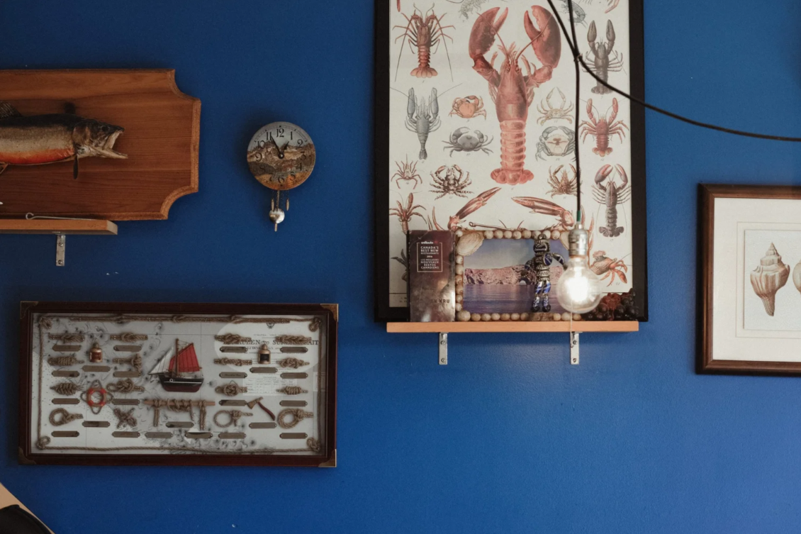 Decorative wall with a framed lobster illustration, a fishing-themed shadow box, a clock, and a framed seashell illustration on a blue wall.