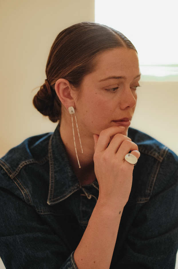 A young woman with brown hair styled in a bun, wearing a denim jacket, with a white earring and ring, resting her chin on her hand in a thoughtful pose indoors.