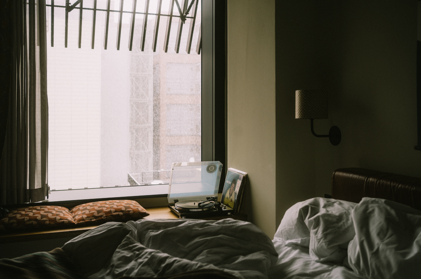 A dimly lit bedroom with an unmade bed, a window with a view of a building and rain outside, and a small table holding a record player, vinyl records, and a magazine.