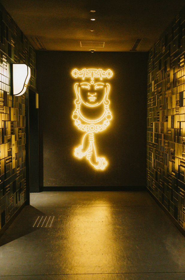 Neon sign of a stylized face with jewelry and a headpiece on a black wall in a hallway.