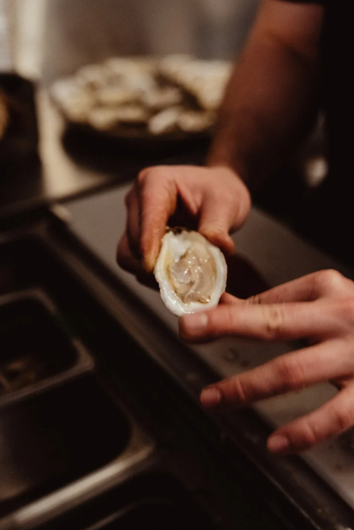 Person holding a raw oyster over a kitchen counter with a bowl of oysters in the background.