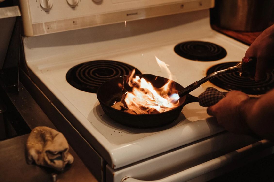 A person is using a torch to set a pan on fire on a white electric stove. The person is holding a cloth in one hand and the torch in the other. The stove has four burners, and the one with the fire is the front right. A cloth or rag is on the counter