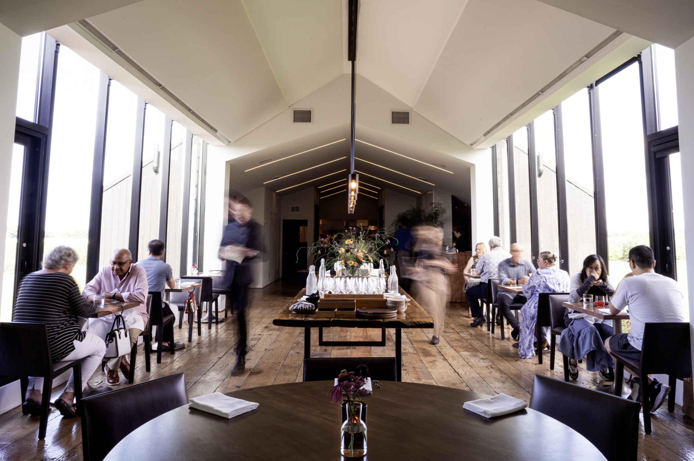 A busy restaurant interior with large windows, high vaulted ceiling, and people dining at tables.