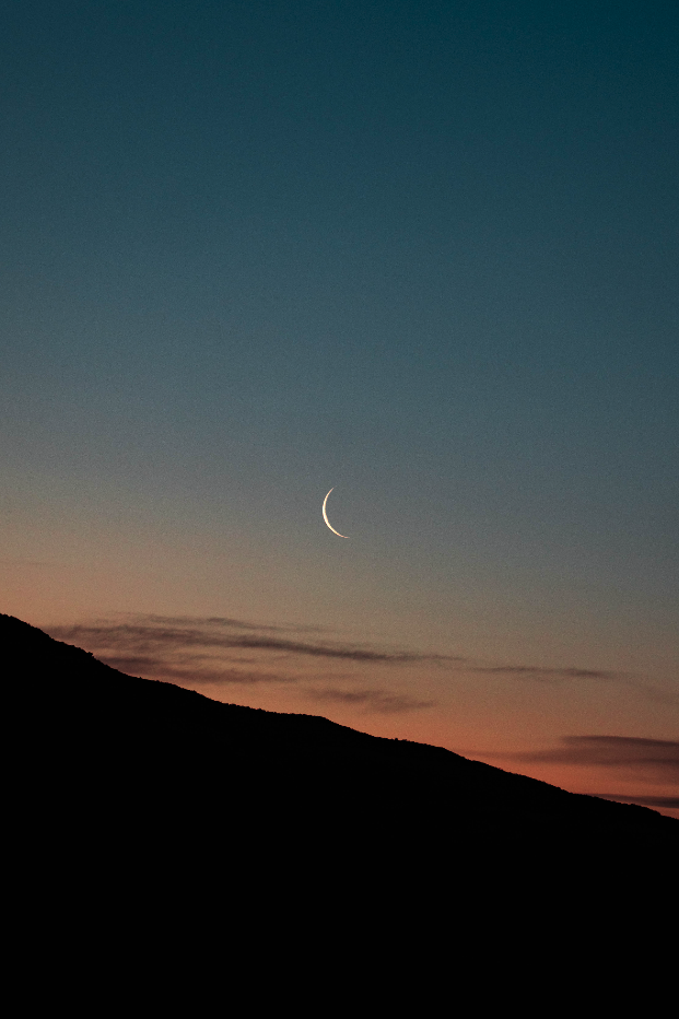 A crescent moon in the sky during dusk, with a silhouette of a mountain range and soft colorful clouds.