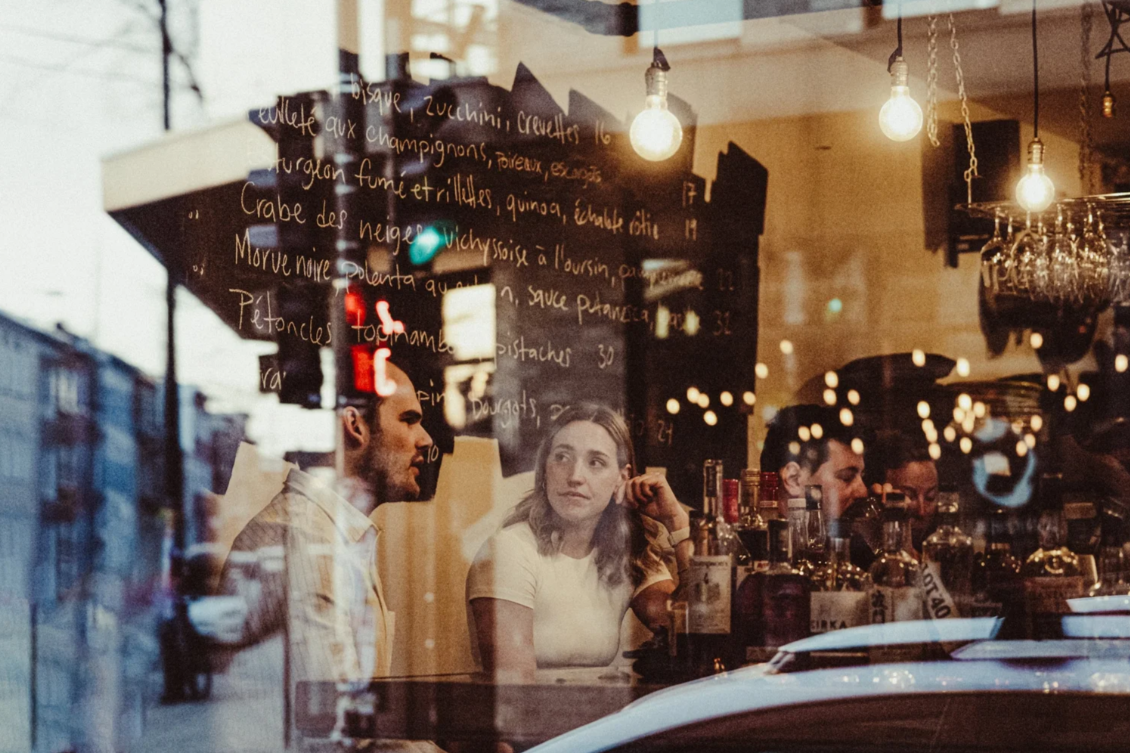People sitting inside a bar or restaurant, seen through a glass window with reflections, bottles on the bar counter, warm lighting, and a menu written on the window.