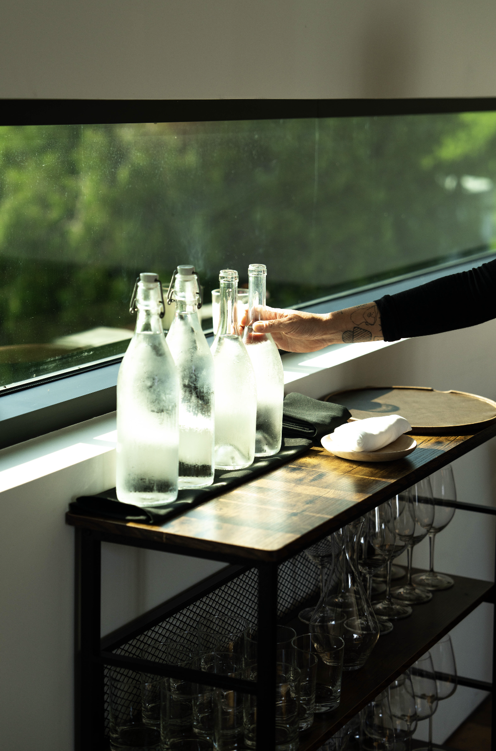 Person arranging glass bottles on a window ledge. The bottles appear to be filled with water or a clear beverage. The scene shows sunlight coming through the window, with a view of greenery outside. There are additional empty glasses and a tray on a 
