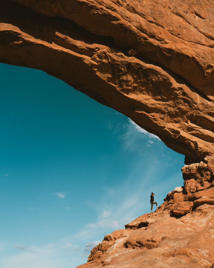 A person standing on a rocky ledge under a natural rock arch formation during daytime.