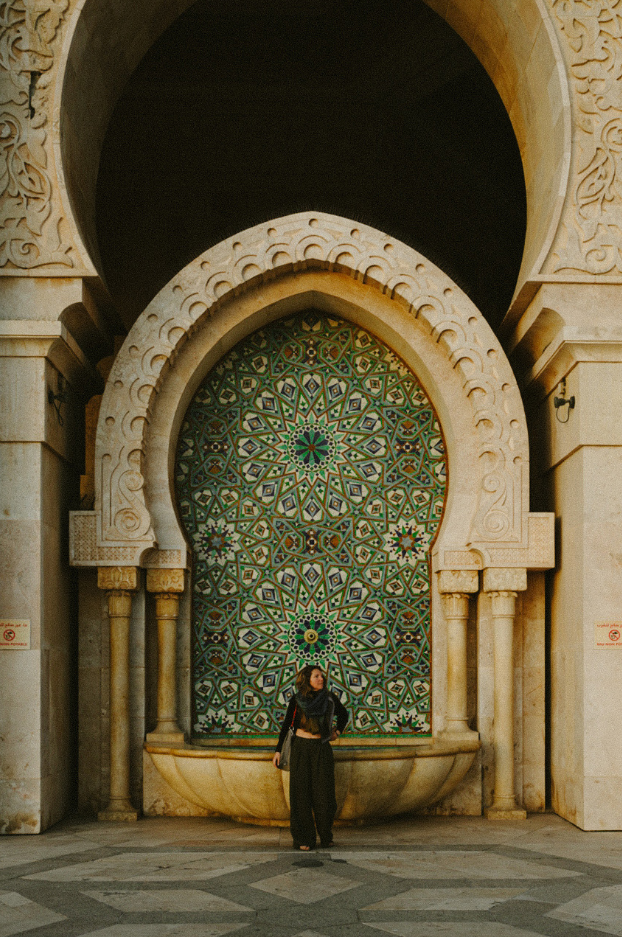 A woman stands in front of a decorative fountain with intricate mosaic tilework and carved stone arches in an ornate architectural setting.