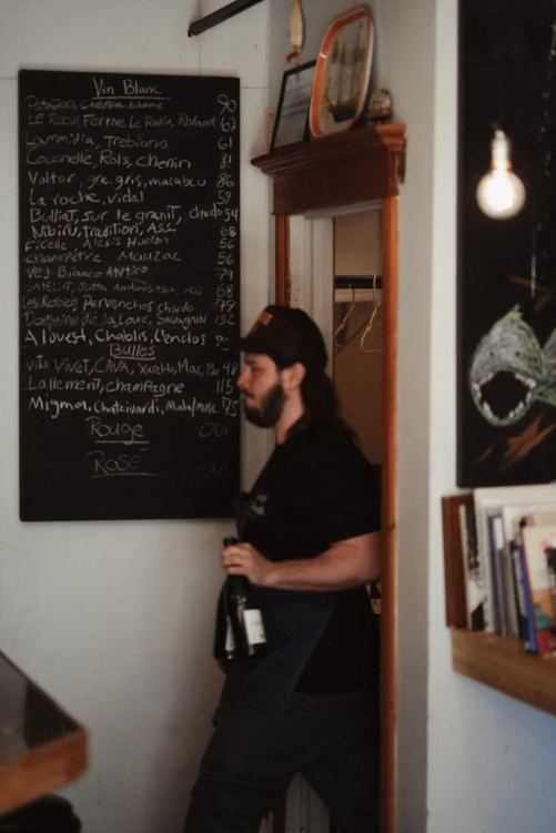 A man with a beard, wearing a black cap and black shirt, holding a wine bottle, standing near a chalkboard menu inside a restaurant or cafe.