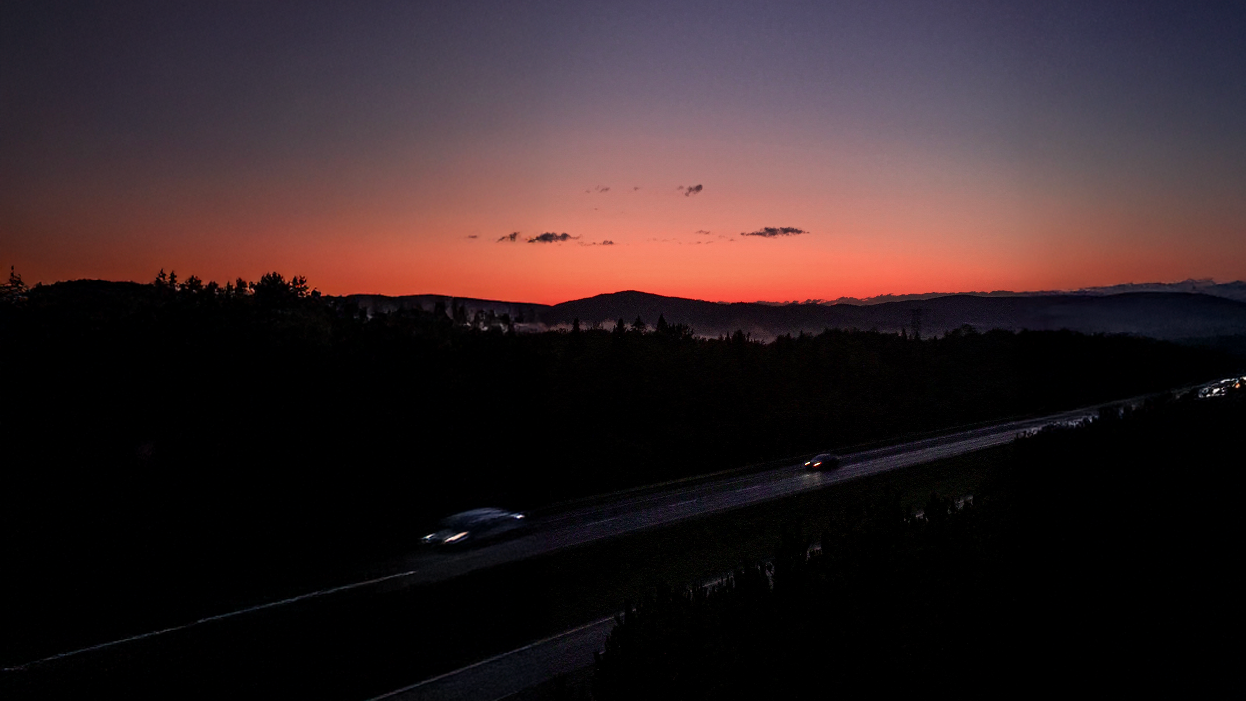 A highway at dusk with cars driving, silhouettes of trees, and mountain ranges in the distance under a colorful sunset sky with pink and purple hues.