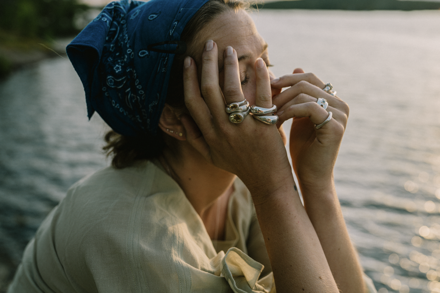 a woman wearing many rings and a blue scarf on her head has her hands in her face over closed eyes in front of a lake