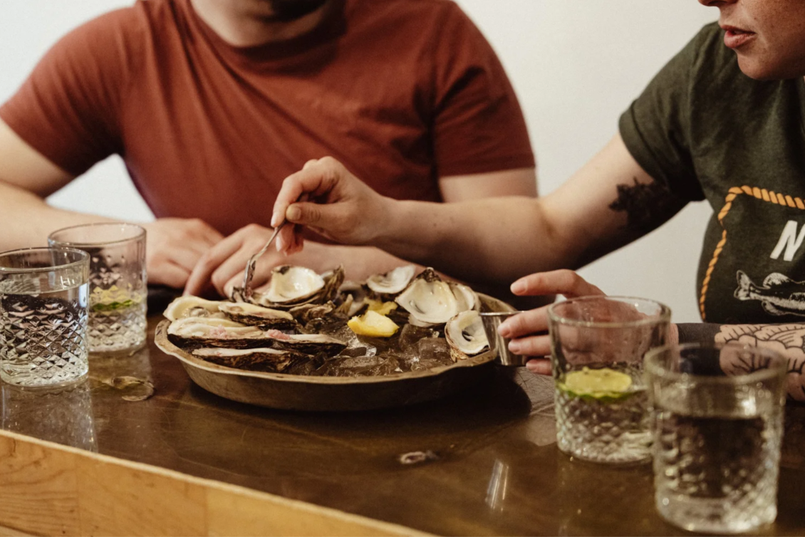 Two people are sharing a plate of shucked oysters on ice with lemon wedges, with glasses of water on a wooden table.
