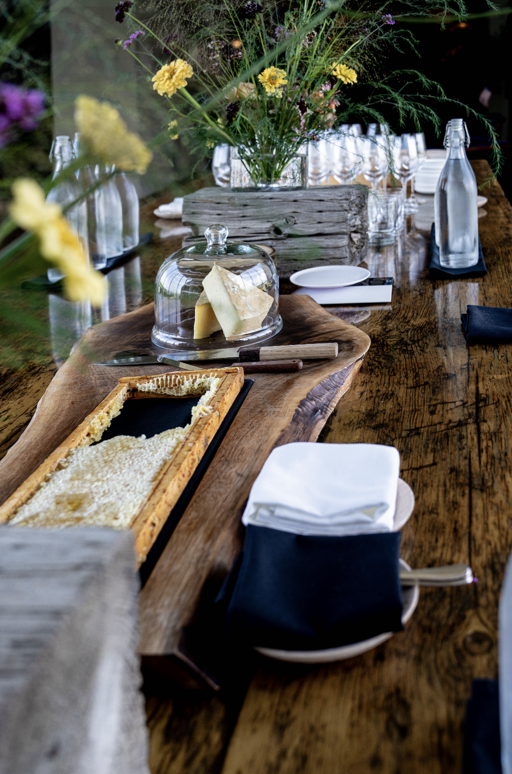 A rustic dining table decorated with a large floral centerpiece, bottles of water, plates, glasses, cheese, and a cheese board, with a wooden table and natural decorations.