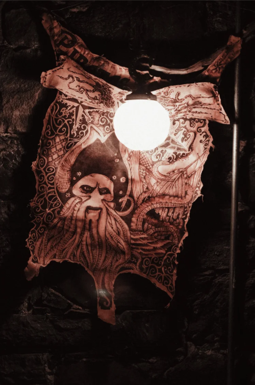 A decorative fabric with a tribal-style design featuring a face with tentacle-like features hanging on a dark brick wall, illuminated by a glowing light bulb above.