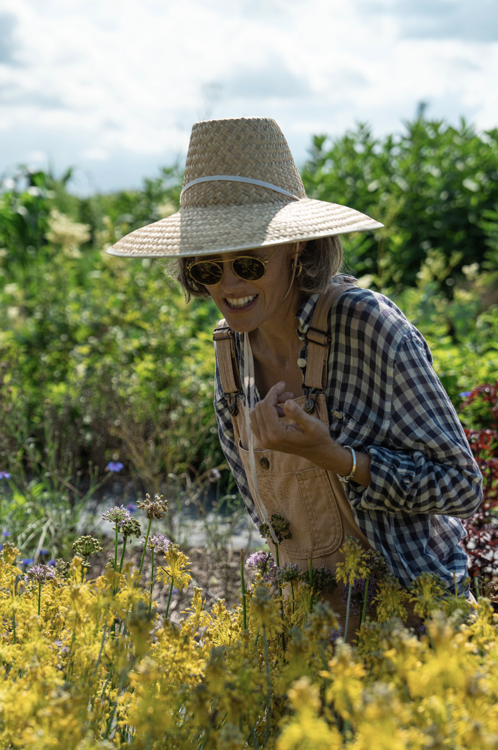 A woman wearing a wide-brimmed straw hat and sunglasses is smiling while working in a flower garden with yellow and purple flowers, wearing a checkered shirt and beige overalls.