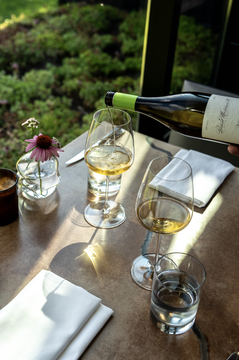 A person pouring white wine into a wine glass at a table set with white napkins, glasses of water, and a small flower arrangement, with greenery visible outside through a window.