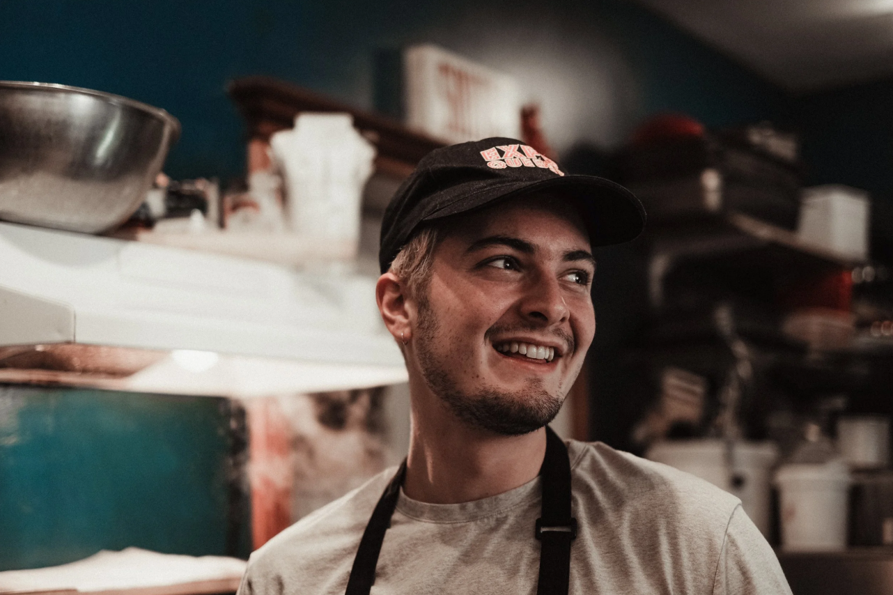 A young man smiling in a kitchen, wearing a black cap and an apron.