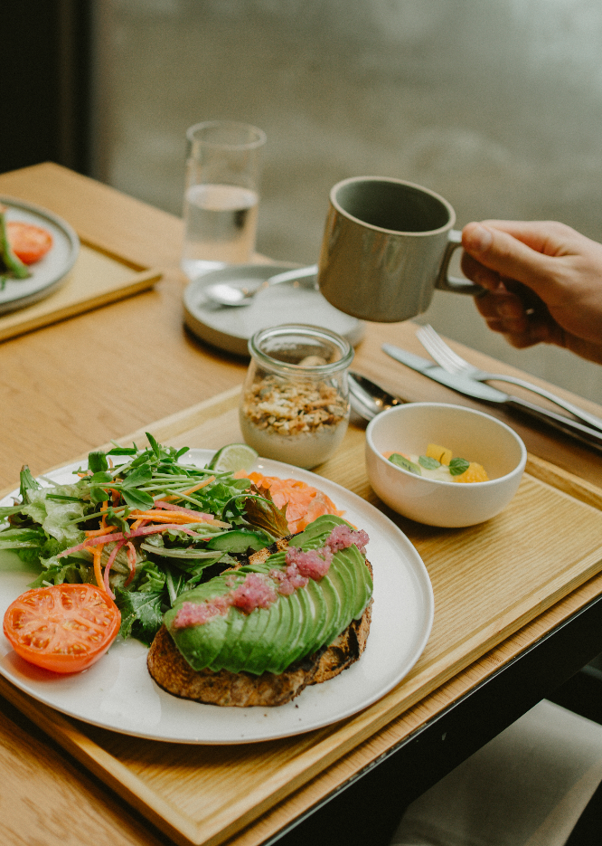 A breakfast tray with a plate of toast topped with sliced avocado and pink spread, a fresh salad with tomatoes and greens, a bowl of fruit, a jar of granola with yogurt, and a cup of coffee, on a wooden table.