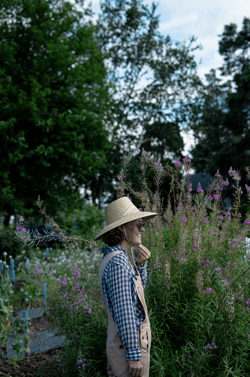 A woman wearing a large straw hat, sunglasses, a plaid shirt, and beige overalls, standing in a garden of tall purple flowers and green trees, smiling and enjoying nature.