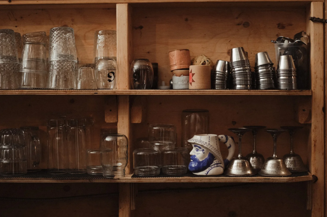Wooden shelving filled with drinking glasses, cups, and barware, including shot glasses, beer mugs, cocktail glasses, and a ceramic mug with a face.