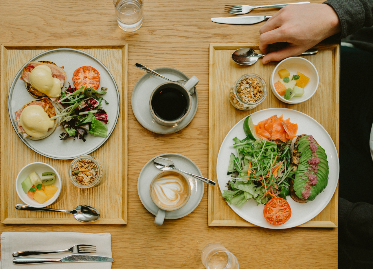 A breakfast setting with two wooden trays holding eggs benedict, a salad, granola, fresh fruit, smoked salmon, avocado toast, and sliced tomatoes. There are cups of coffee, a glass of water, and utensils.