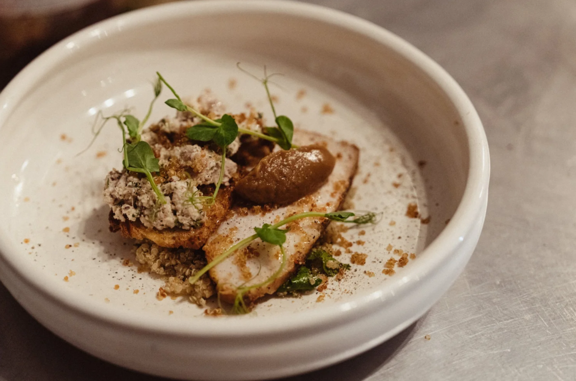 A white bowl of dessert with ice cream, a piece of cake or biscuit, a dollop of chocolate or caramel sauce, microgreens, and powdered spices.