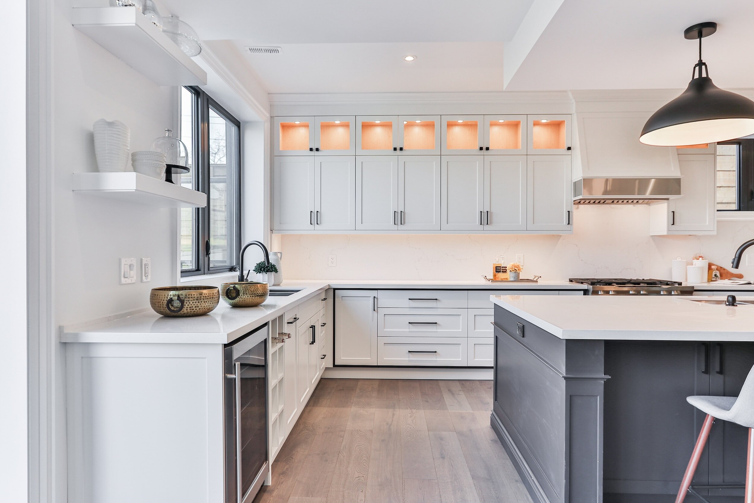 Modern kitchen with white cabinets, black island, pendant light, and wooden flooring. Shelves with decorative items, built-in oven, and large windows for natural light.