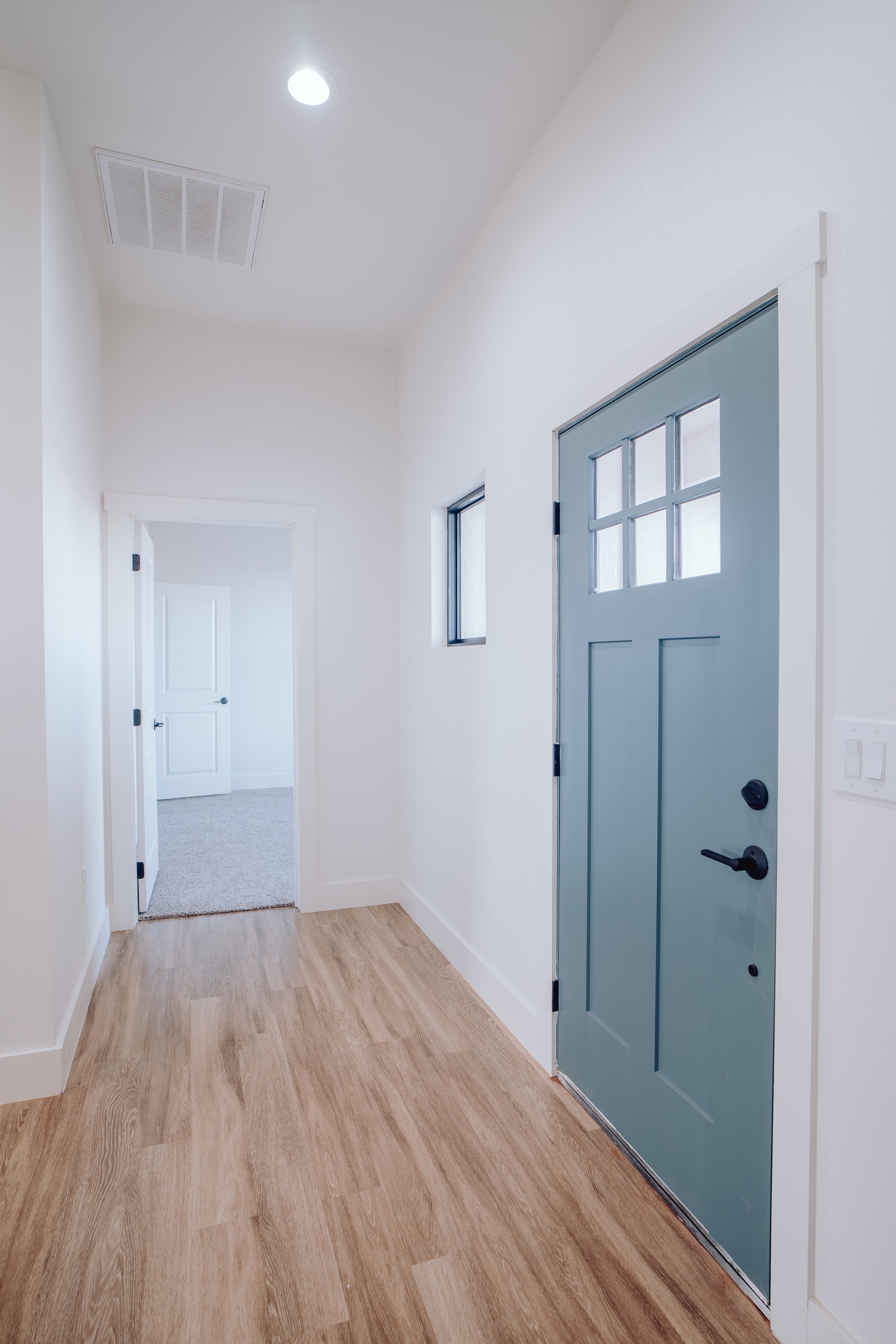Interior hallway with hardwood flooring, light blue front door with glass panels, white walls, square ceiling light, and a doorway leading to a carpeted room.