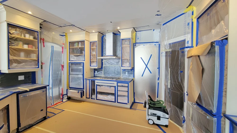 Kitchen prepared for refinishing with protective masking over appliances and surrounding surfaces.