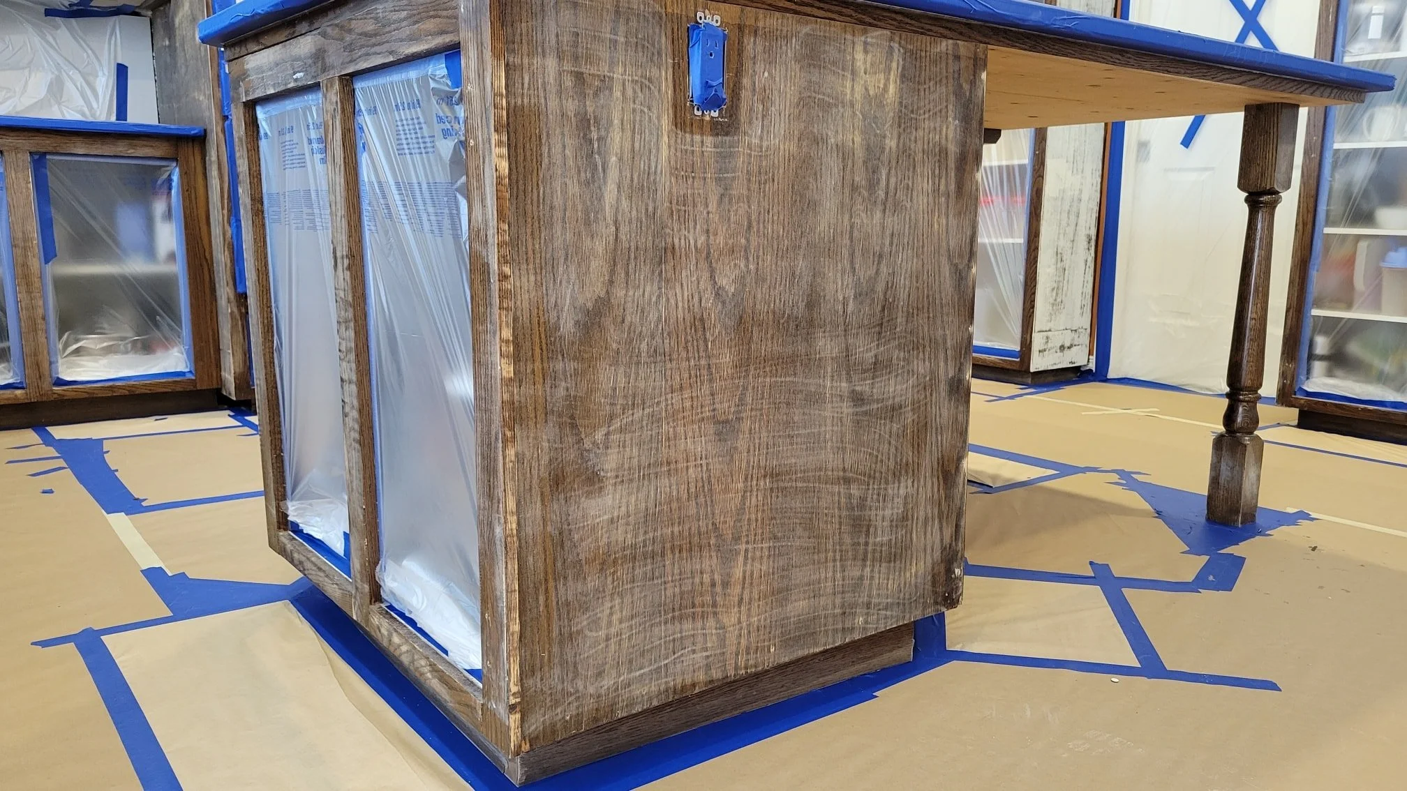 Wooden kitchen island under construction, surrounded by protective tape and plastic, with unfinished cabinetry.