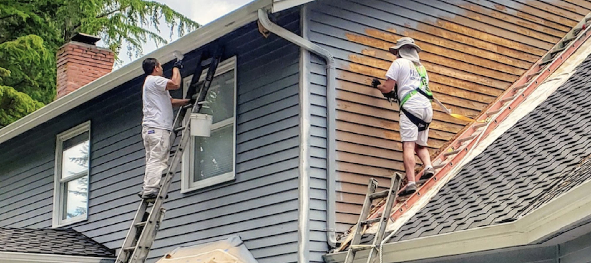 Two workers on ladders replacing or cleaning the siding and roof of a house. One worker is on the higher ladder close to the roof, and the other is on a lower ladder near the edge of the roof.