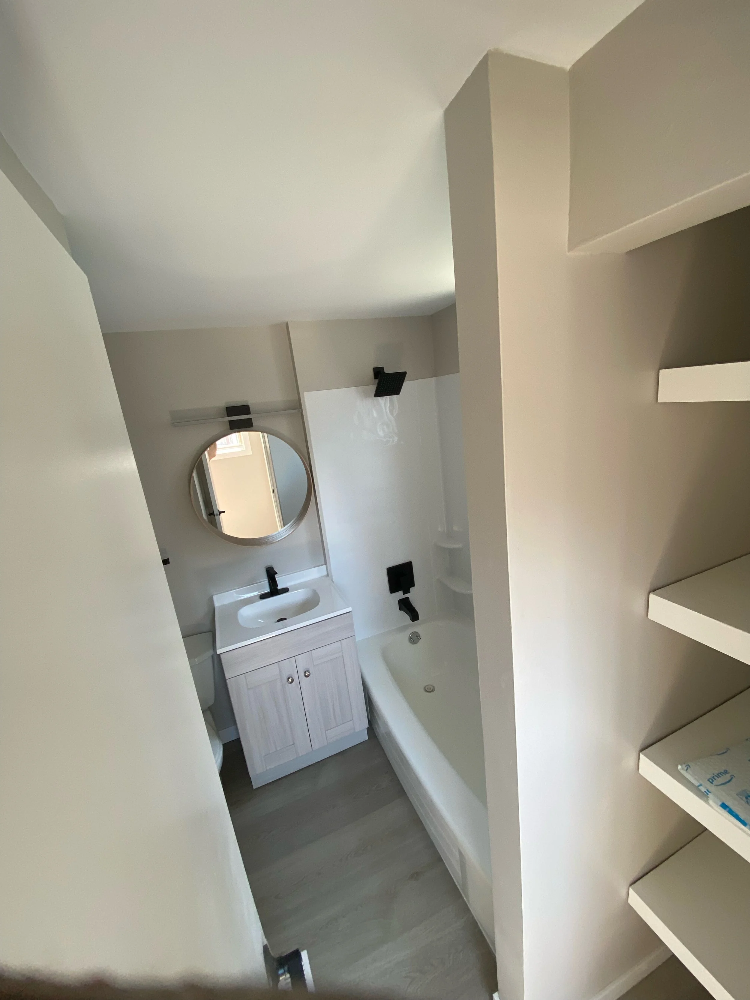 Small bathroom with white vanity, round mirror, bathtub, and built-in shelves.