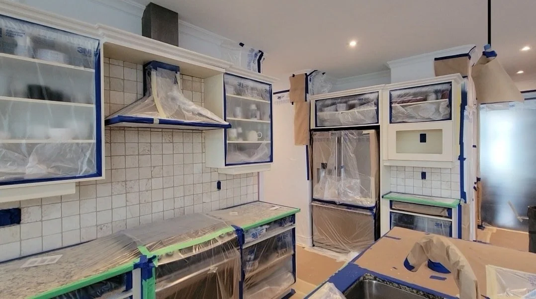 Kitchen under renovation with cabinets and appliances covered in protective plastic sheeting, preparing for painting or construction work.