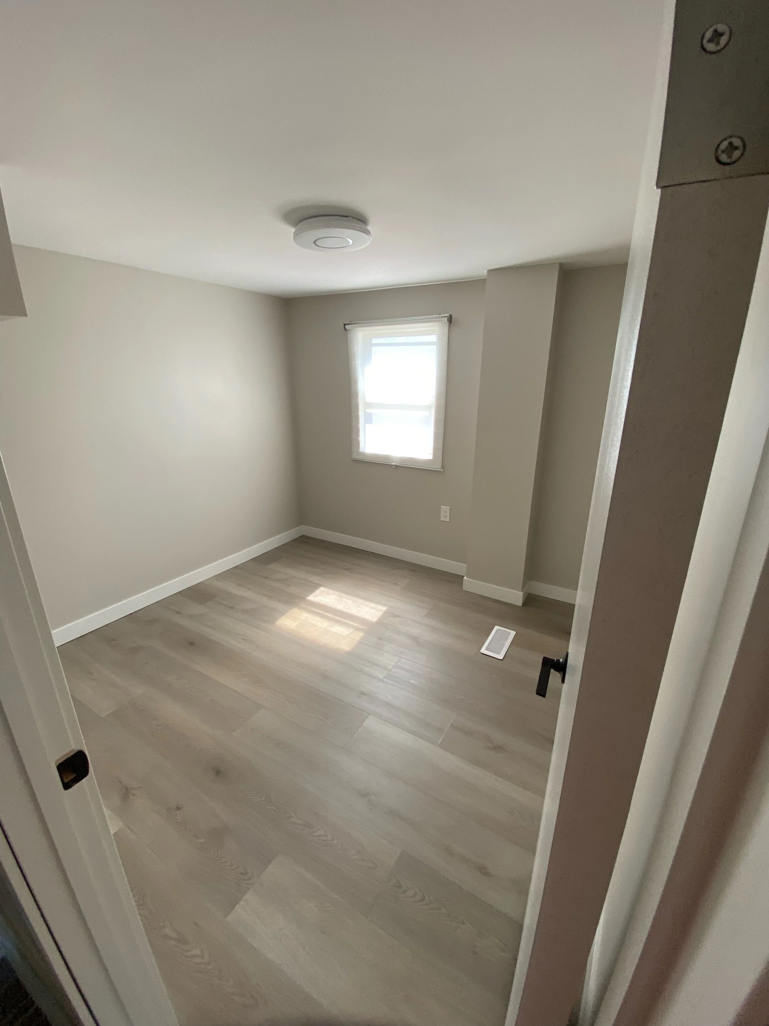 Empty room with light wood flooring, beige walls, a window, and a ceiling fixture.