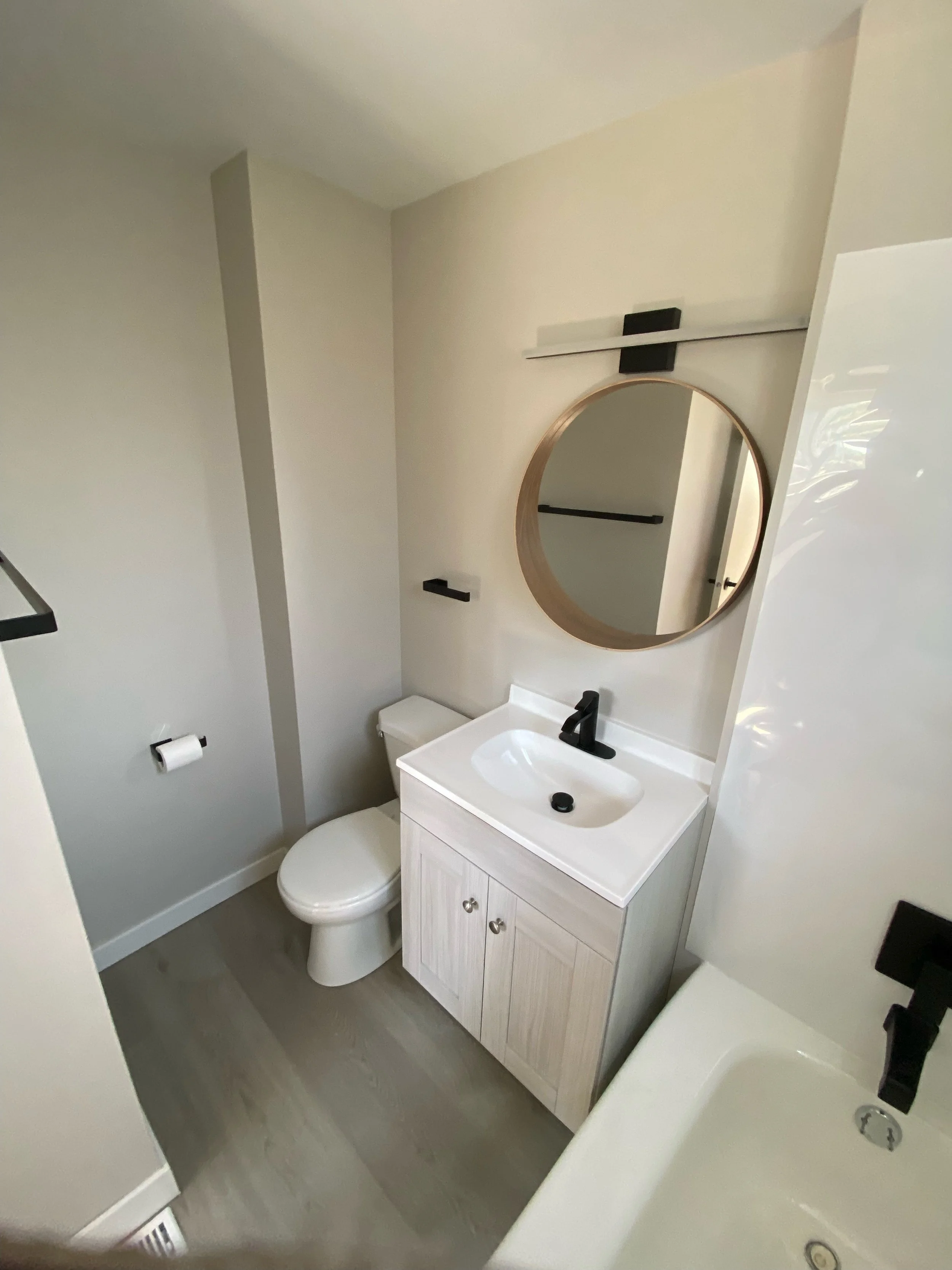 Modern bathroom with light wood vanity, round mirror, toilet, black fixtures, and beige walls.