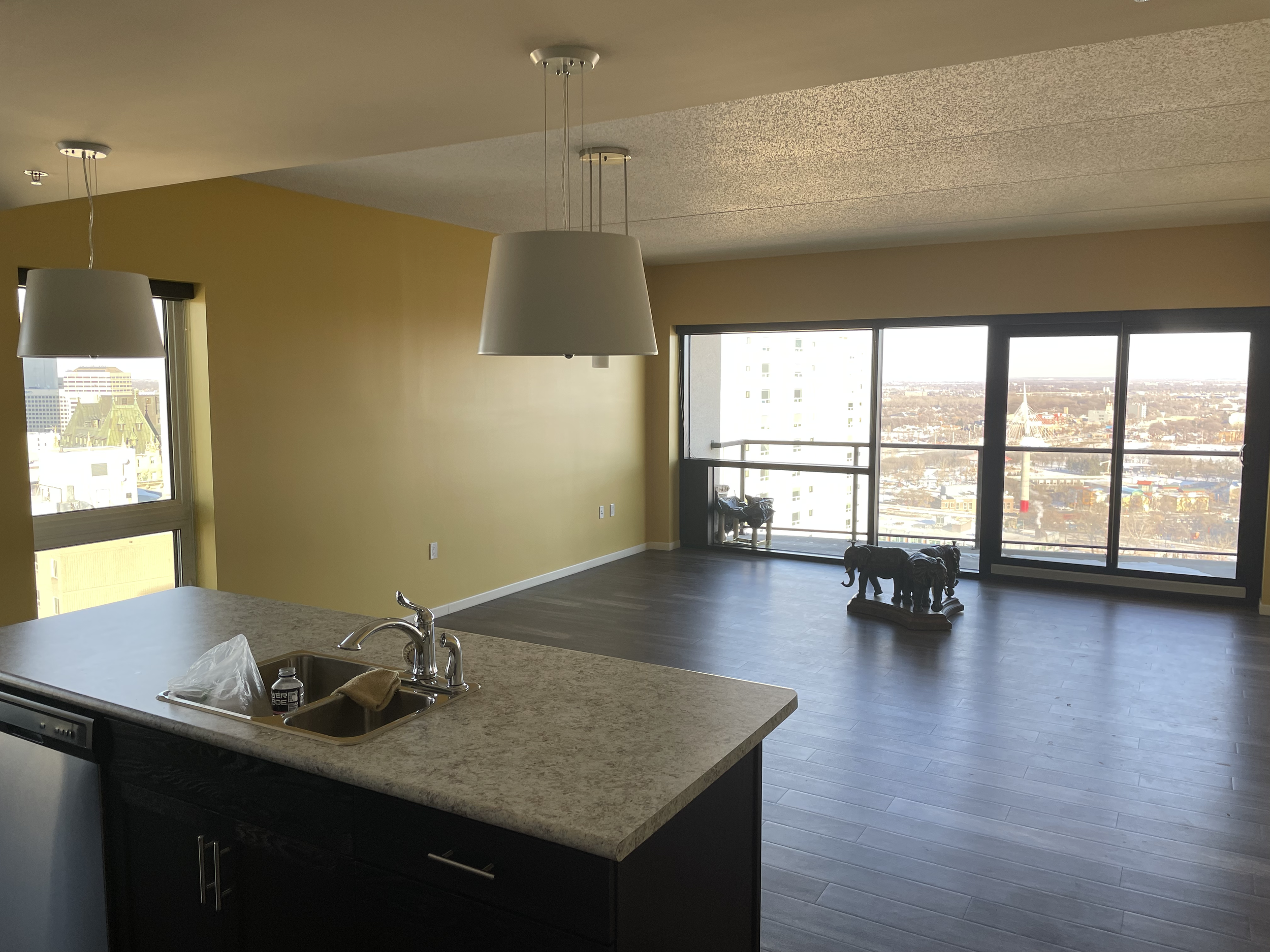 Modern apartment interior with large windows, a kitchen island, hanging pendant lights, and decorative elephant figurines on the floor.