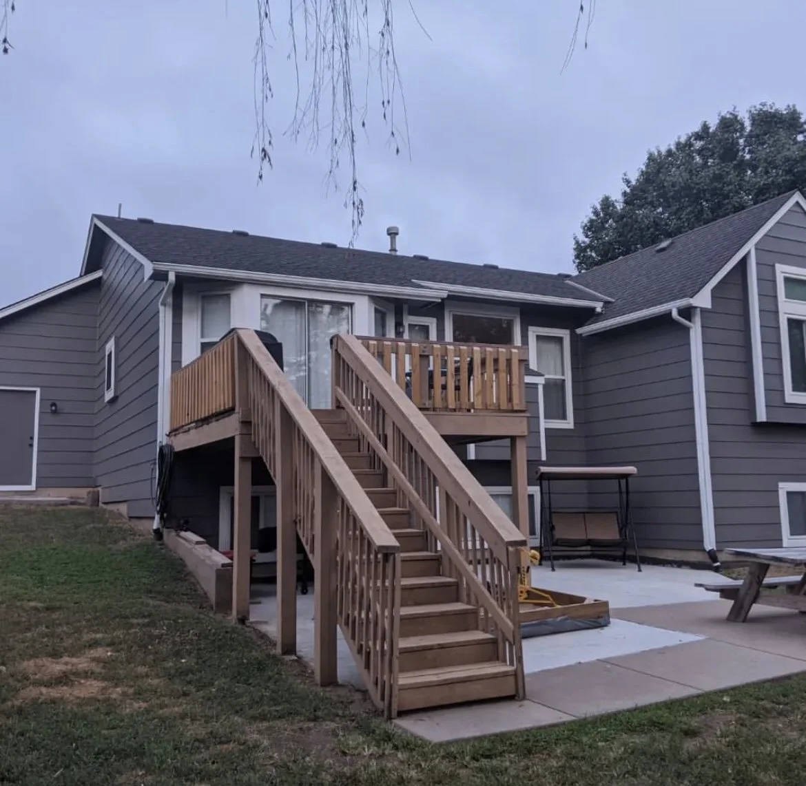 Residential backyard with a wooden deck and stairs, gray house exterior, grass lawn, and patio area with bench.