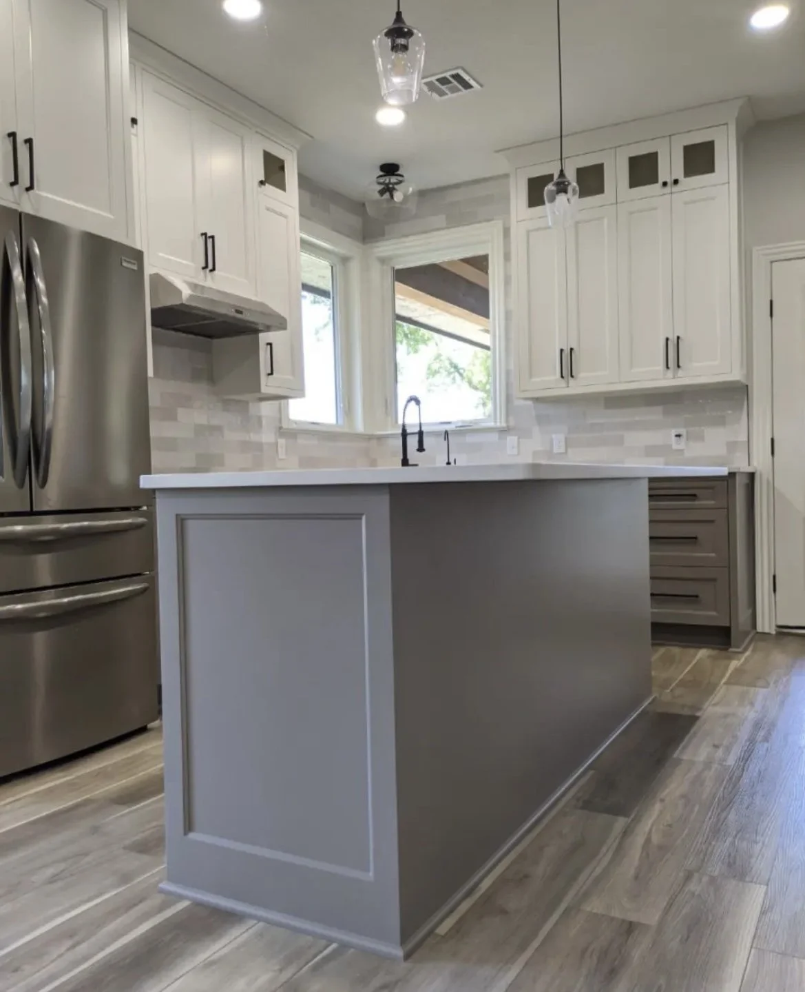 Modern kitchen with gray island, stainless steel refrigerator, white cabinets, pendant lights, and wood flooring.