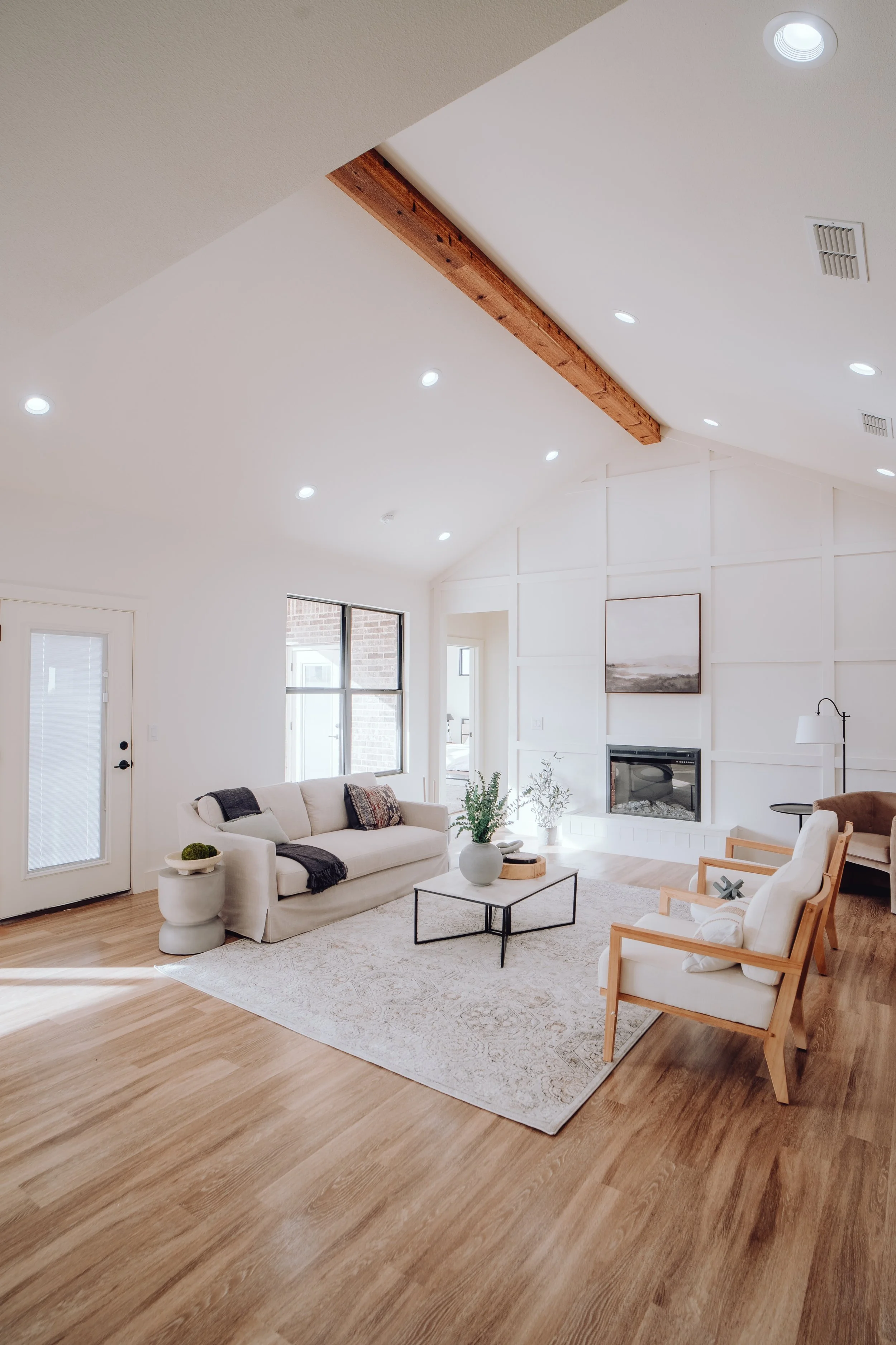 Minimalist living room with vaulted ceiling, light wood flooring, and a beige sofa set. Features a modern coffee table, wall art, white armchairs, and a large window.