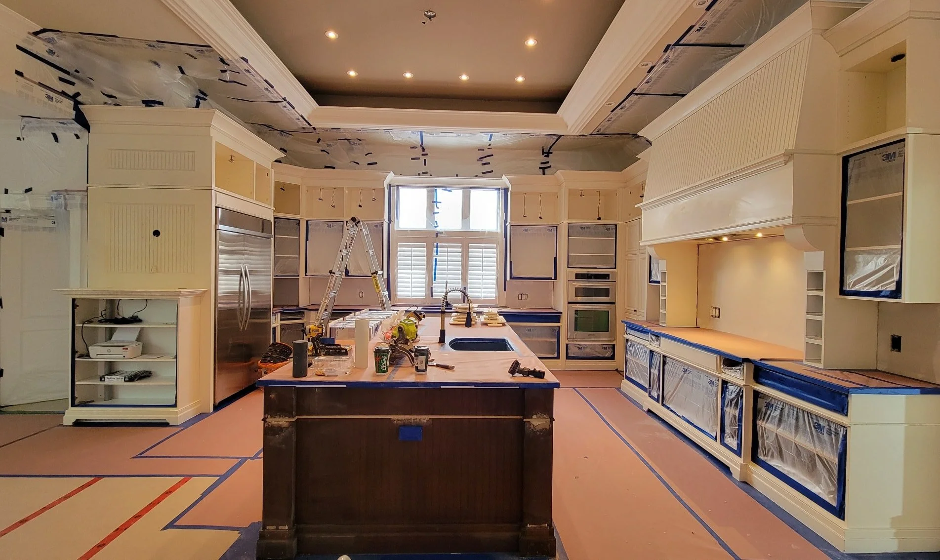 Modern kitchen under renovation with white cabinetry, large island, stainless steel appliances, and covered surfaces. A ladder and tools are present.