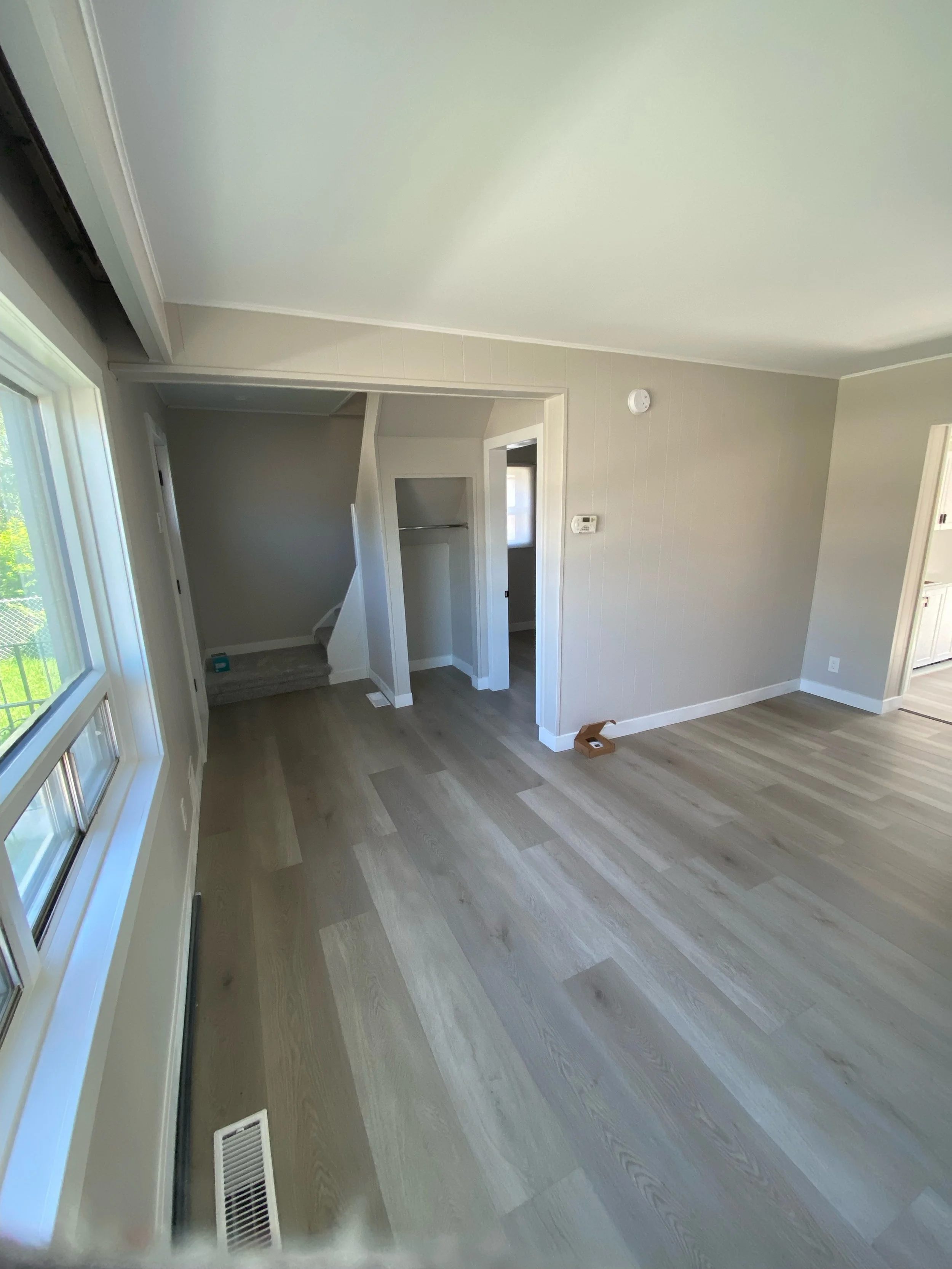 Interior of an empty room with light wood flooring, gray walls, and large windows. There is a small closet and an opening leading to another area.