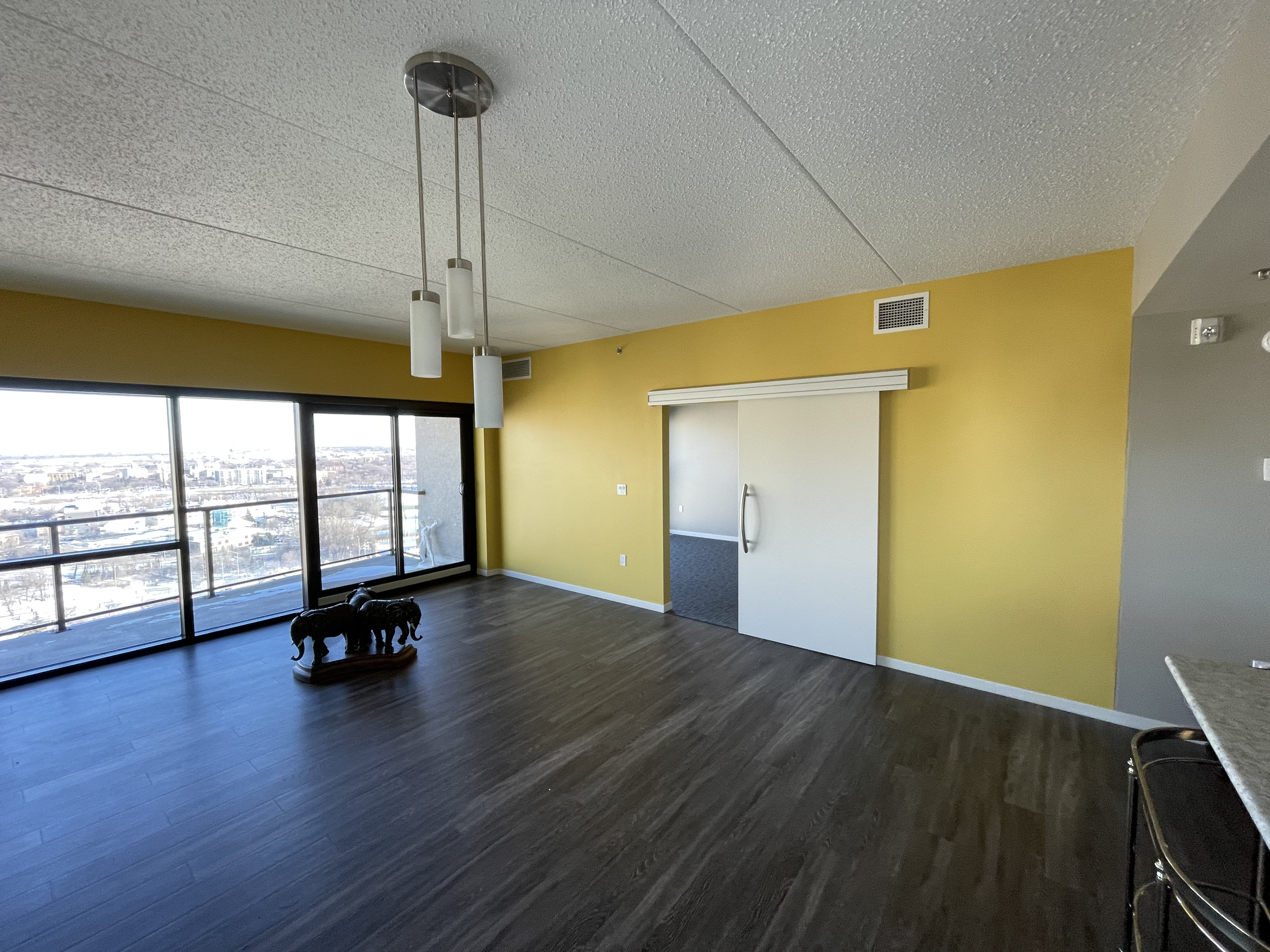 Spacious apartment interior with yellow painted walls, large sliding glass doors leading to a balcony with city view, wooden flooring, minimalist pendant lighting, and decorative elephant statues on the floor.