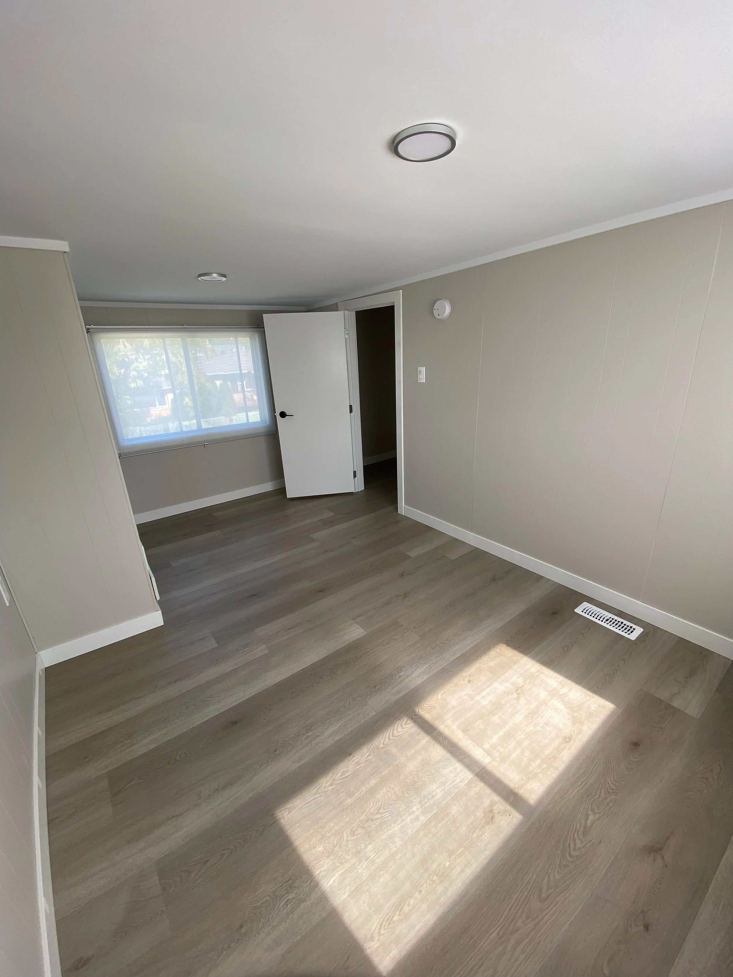 Empty room with wooden floors, beige walls, a white ceiling, and a window with a view outside. The door is open, revealing an adjoining space. Ceiling lights and a wall vent are visible.