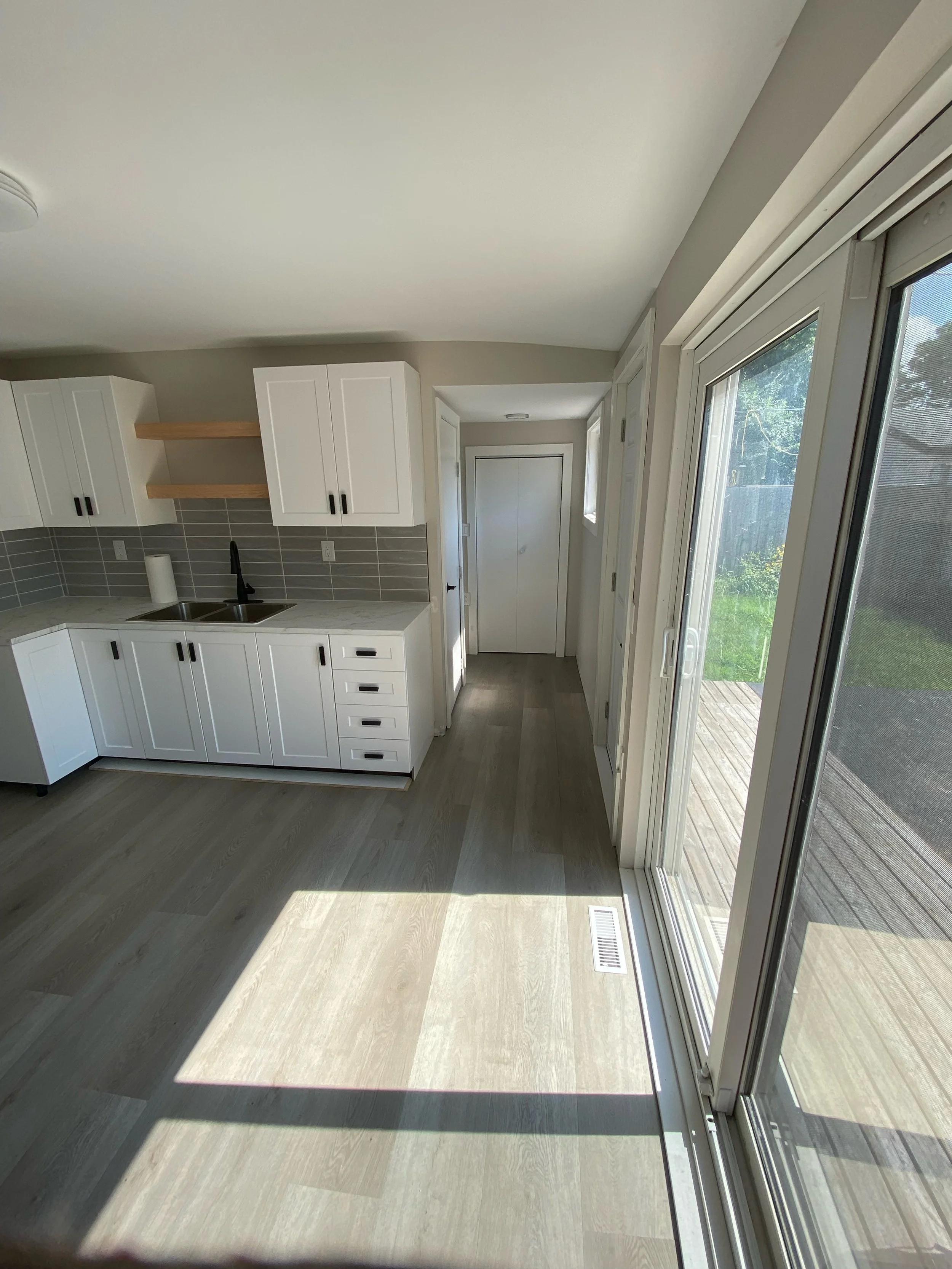 Modern kitchen interior with white cabinets, wood shelving, and sliding glass door leading to outdoor space.