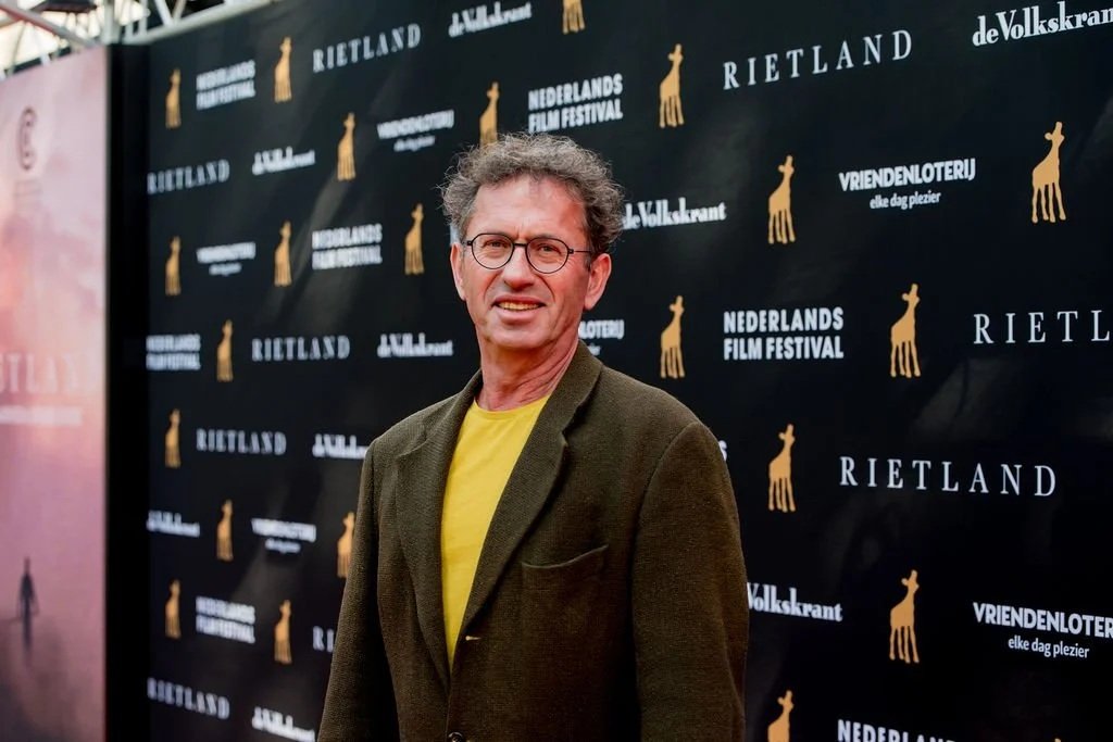 Man met bril staat op de Rode Loper van het Nederlands Film Festival. Hij draagt een donker colbert met een okergeel t-shirt. 