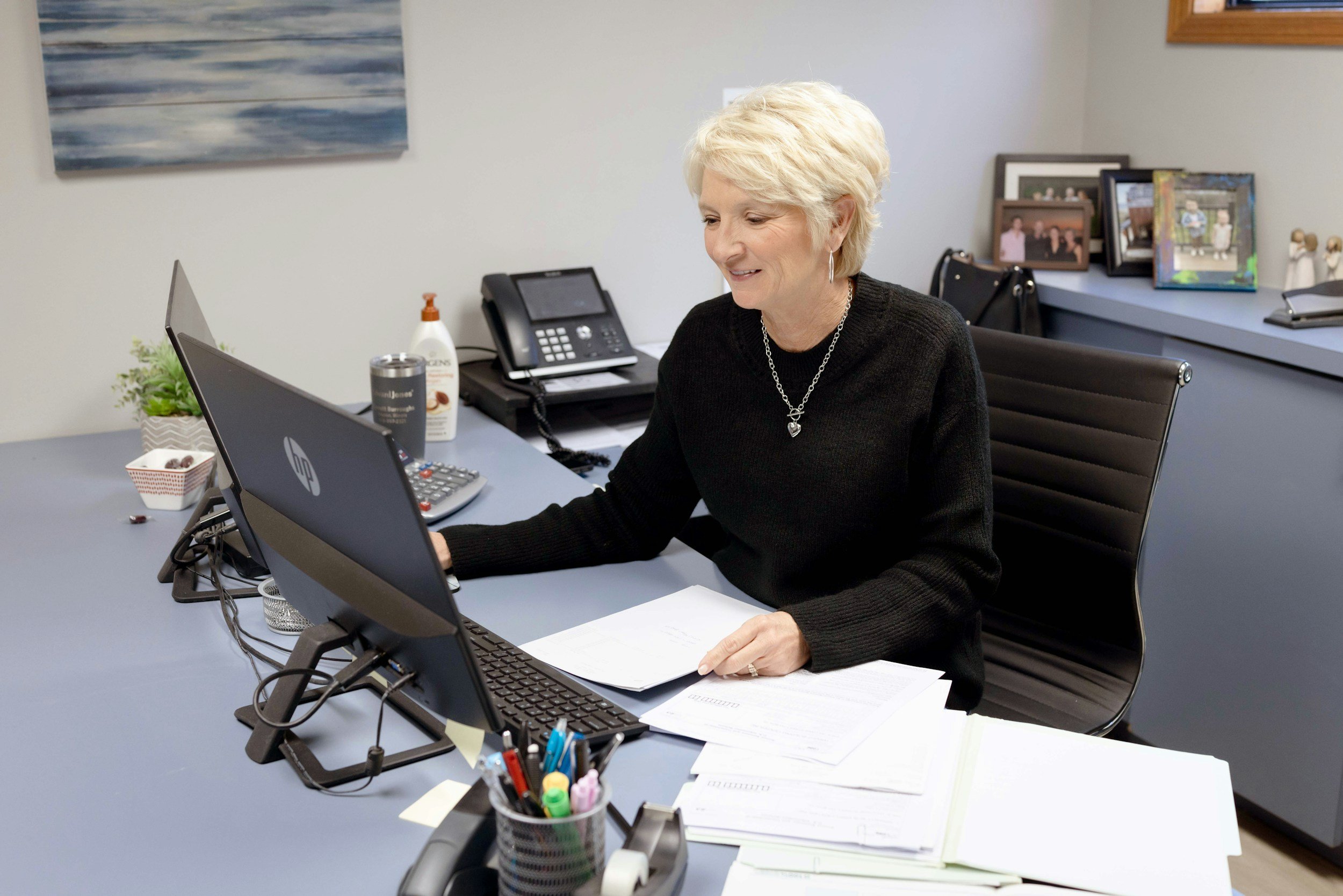 An elderly woman with short blonde hair working at her office desk, looking at a computer monitor, with papers and office supplies around her.