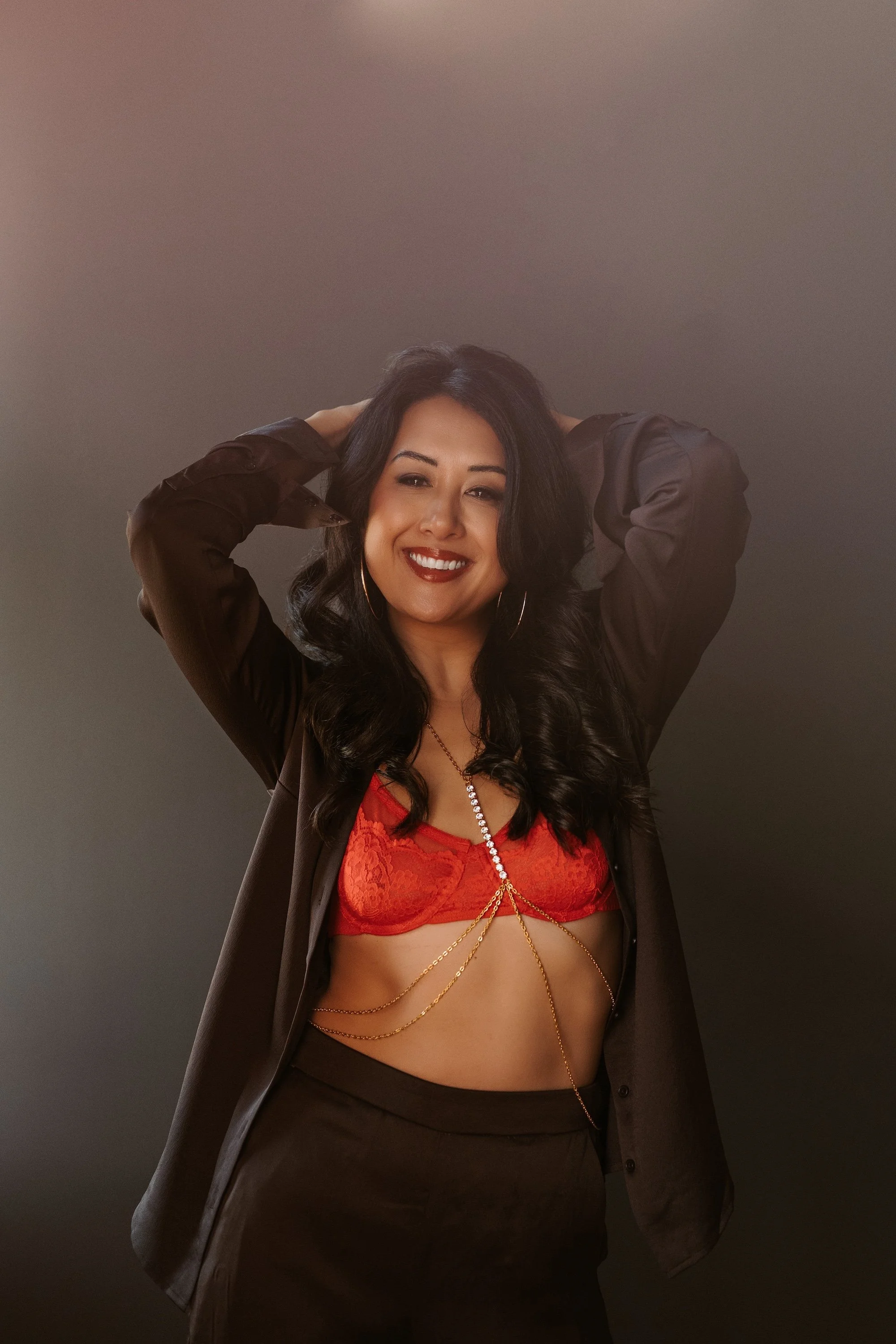 A woman with long wavy hair smiling, wearing a black blazer over a red lace bra, accessorized with gold jewelry, standing against a plain dark background.