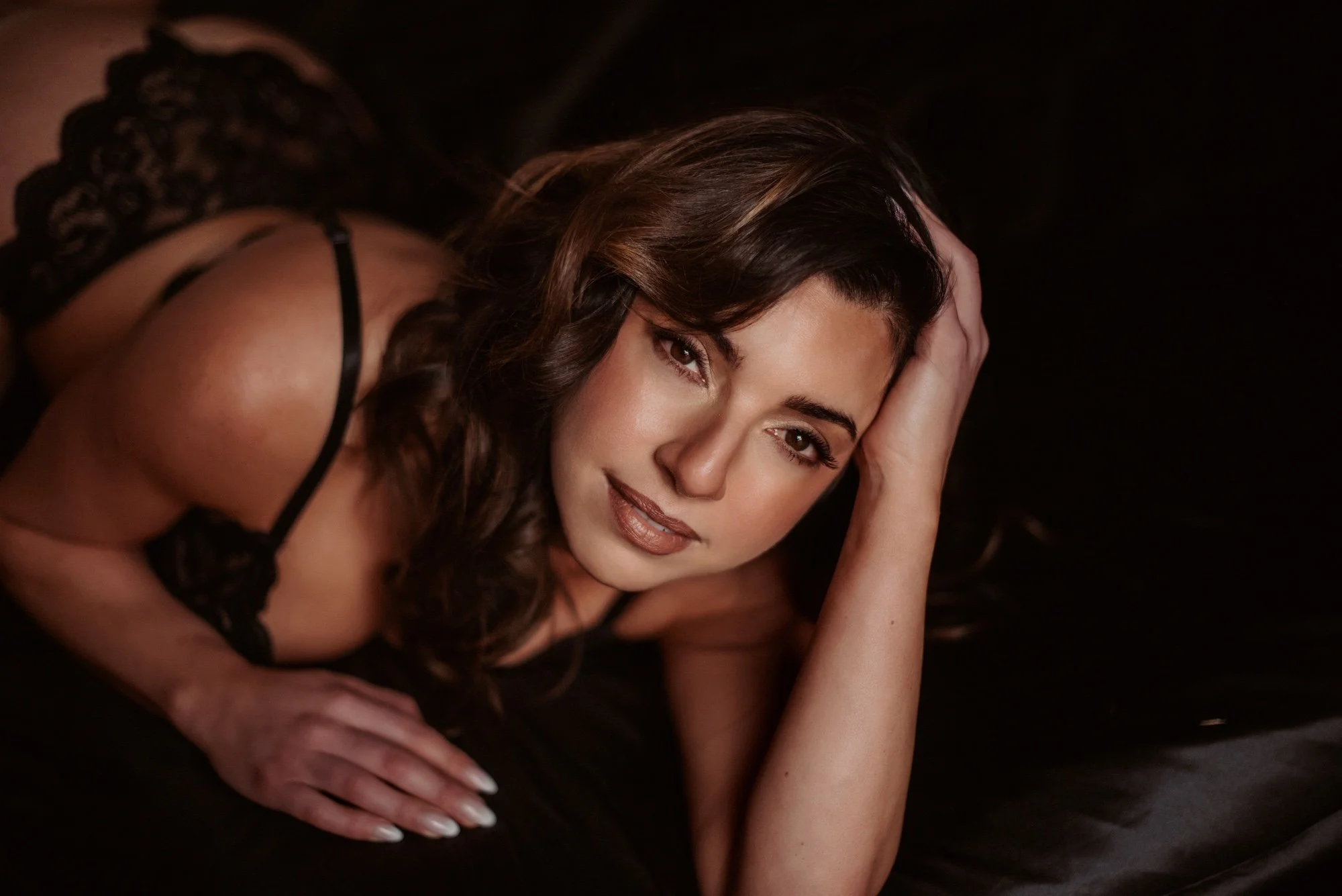Woman with long brown hair lying on her side, resting her head on her hand, wearing black lace lingerie, against a dark background.