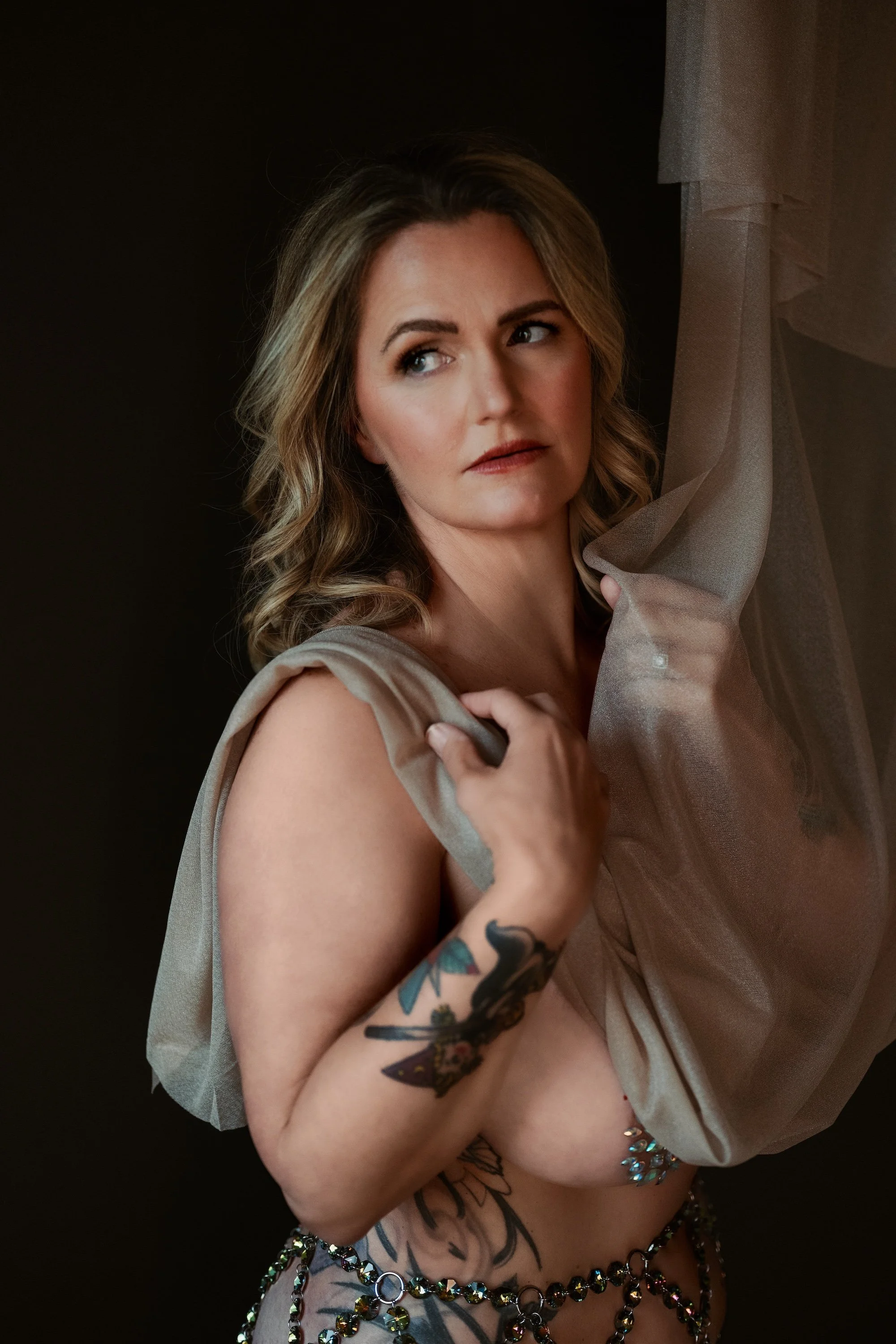 A woman with long wavy blonde hair, gazing to her left, partially covered with sheer beige fabric, standing against a dark background, wearing a chain of multicolored beads around her waist, tattoos on her left arm and side, and multiple bracelets on