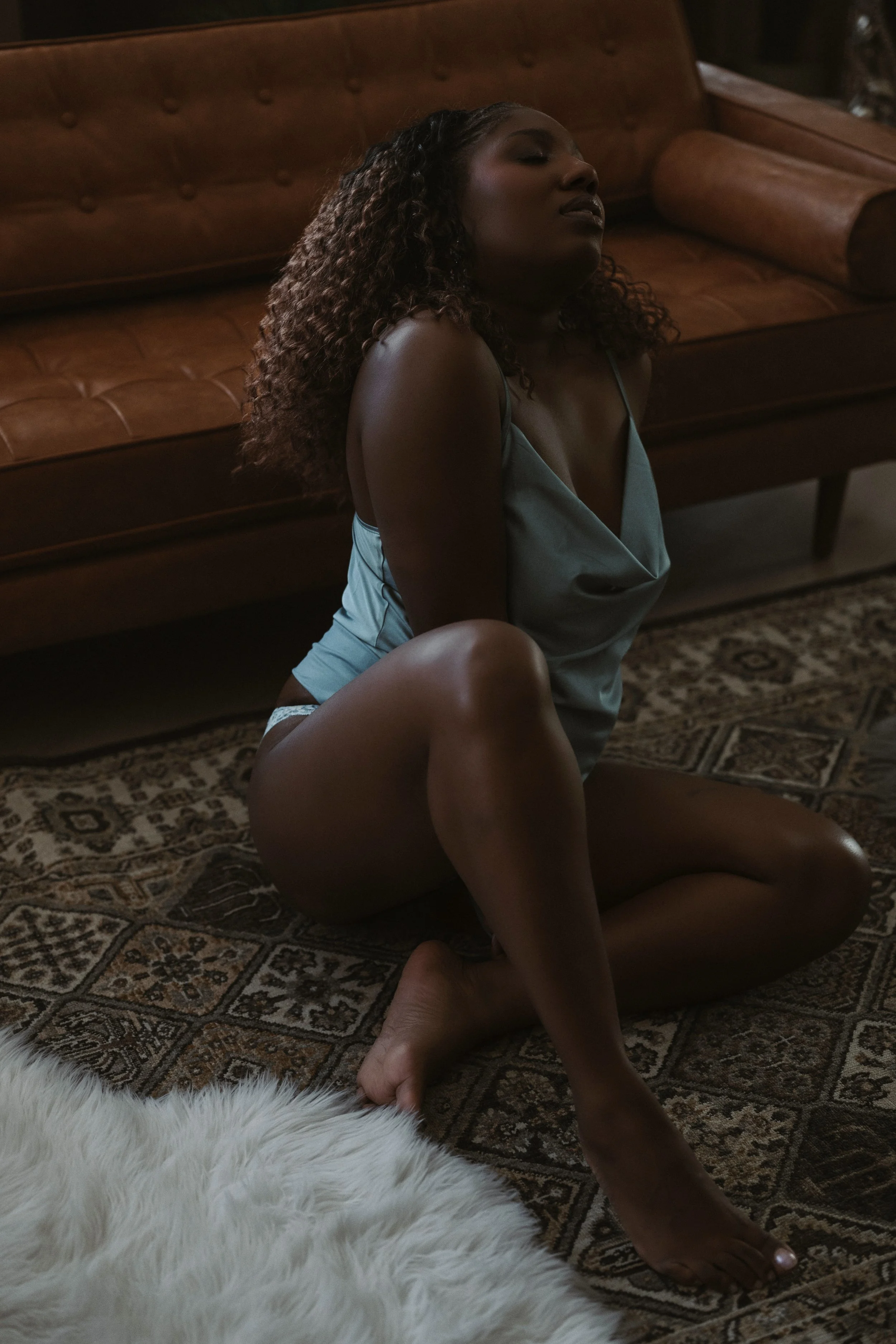 A woman with curly hair is sitting on the floor with her eyes closed, wearing a light blue satin slip, next to a brown leather sofa and a patterned rug, with a white furry rug in the foreground.